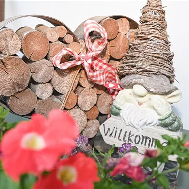 A decorative heart made of wooden logs with a red gift ribbon and a small, friendly garden gnome figure featuring the inscription "Welcome". Colorful flowers frame the scene.