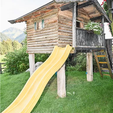 A wooden playhouse with a yellow slide stands on a grassy field. In the background, mountains and plants are visible.