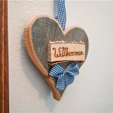 A heart-shaped welcome sign made of wood, adorned with a blue checked ribbon. The sign hangs on a wall and exudes coziness.