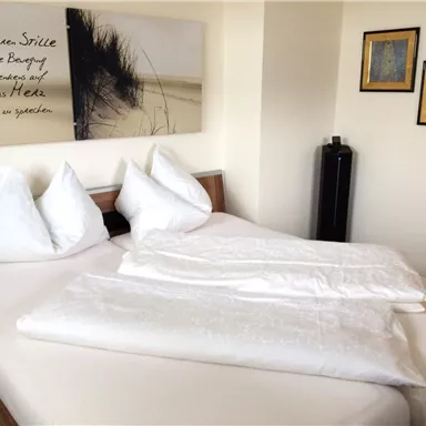 A cozy bedroom with a double bed and white pillows. Pictures hang on the wall along with a lettering that conveys peace and relaxation.