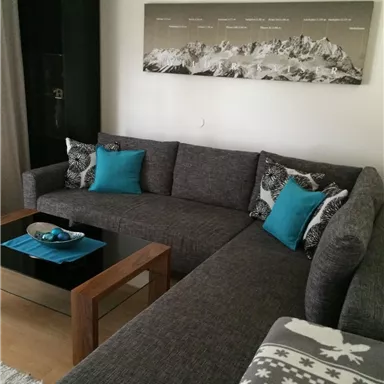 A modern living room with a gray couch and cushions in turquoise. The wooden coffee table and a wall decoration featuring a mountain motif complete the picture.