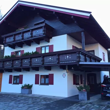 A traditional alpine house with a wooden balcony railing. The facade is white with red shutters, surrounded by a paved driveway and plants.