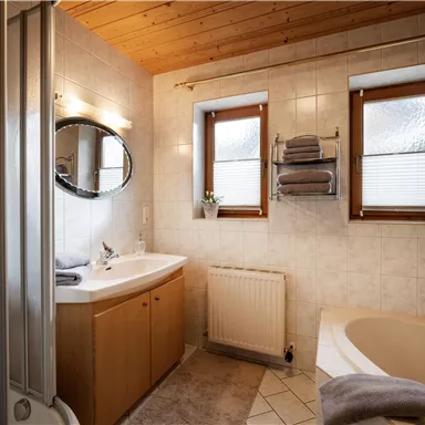 A modern bathroom with a wooden ceiling and bright tiles. There is a bathtub, a sink, and windows with pleats.