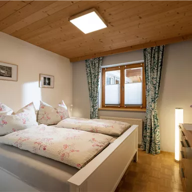 A cozy bedroom with a large bed and floral cushions. It has wooden ceilings and windows with curtains.
