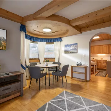 A cozy living area with a dining space, chairs, and a wooden table. The kitchen in the background is well-equipped, and there are natural wood details in the room.