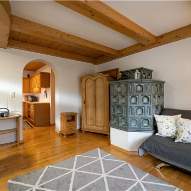 A cozy living space with wooden ceilings and a traditional stove. The room is bright and inviting, with a gray carpet and colorful cushions on the sofa.