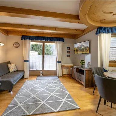 A cozy living room with a wooden beam ceiling and a large window. The room is modernly furnished with a couch, a table, and a carpet.