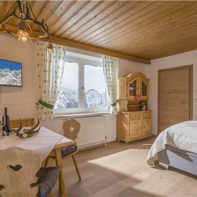 A cozy room with a wooden ceiling and modern television. There is a dining table with chairs, a comfortable sofa bed, and large windows with mountain views.