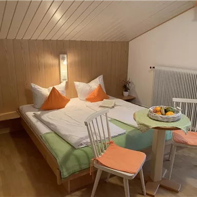 A cozy bedroom with a double bed and orange cushions. Next to the bed is a table with two chairs and a bowl of fruit.