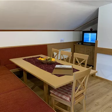 A cozy seating area with a large table and chairs. In the background, there is a television and an appealing decoration.