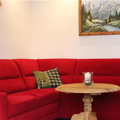 A cozy living room with a red couch and a round wooden table. A landscape painting hangs on the wall.