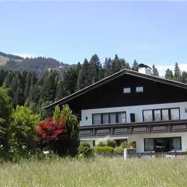 A large house surrounded by green meadows and trees. In the background, gentle hills and a blue sky are visible.