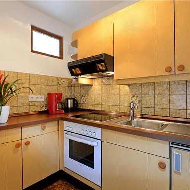 A modern kitchen with wood-colored cabinets and a countertop. In the background, there are a stove, a sink, and a decorative plant visible.