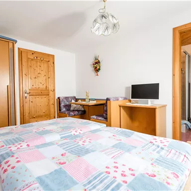 A cozy bedroom with a double bed and a wooden wardrobe. In the background, a small dining area and a bathroom are visible.