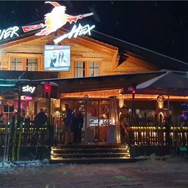 A cozy mountain cabin with an inviting outdoor area, illuminated by colorful lights. Many people enjoy the atmosphere in front of the cabin.