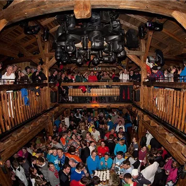 A lively event in a wooden building with many people gathering. There is an upper and a lower floor, both are well attended.