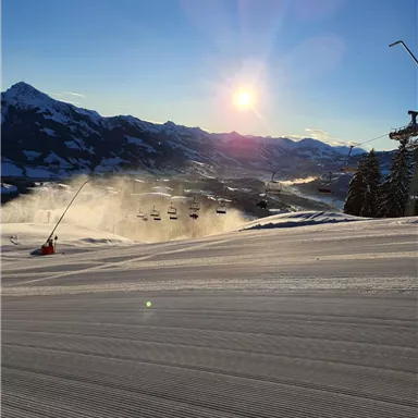 Eine verschneite Landschaft mit frisch präparierten Pisten und einem Sessellift. Im Hintergrund strahlt die Sonne über den Bergen.