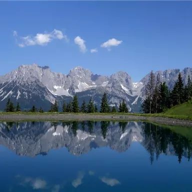A clear lake with a calm water surface reflecting the majestic mountains and the blue sky. Surrounded by green trees and an idyllic landscape.