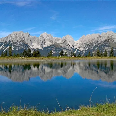 Eine atemberaubende Berglandschaft spiegelt sich im ruhigen Wasser eines Sees. Der klare Himmel und die grünen Wiesen ziehen den Betrachter in die Natur.