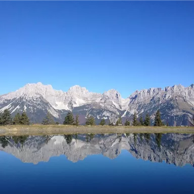 A clear lake in the mountains reflects the impressive peaks. The sky is blue and cloudless.