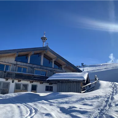 Ein gemütliches Holzhaus im Schnee unter klarem blauen Himmel. Die Umgebung ist von einer dicken Schneedecke bedeckt.
