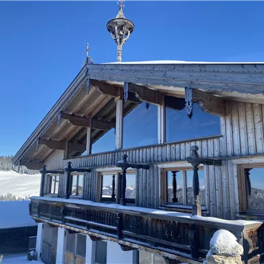 Ein charmantes, holzverkleidetes Chalet im Schnee mit großen Fenstern und einem traditionellen Dach. Die hügelige Winterlandschaft im Hintergrund vermittelt eine ruhige Atmosphäre.