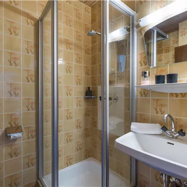 Ein kleines Badezimmer mit einer Dusche und einem Waschbecken. Die Wände sind mit einer hellen Fliesenoptik dekoriert.