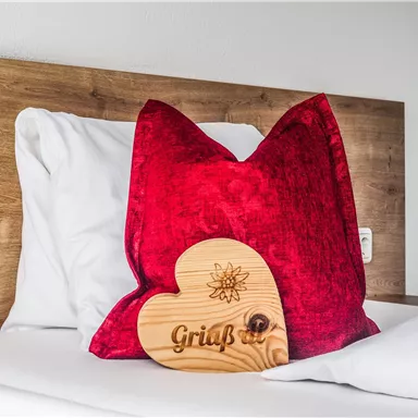 Ein rotes Kissen auf einem Bett mit einem Holzschild in Herzform. Das Schild trägt die Aufschrift „Gruß“.