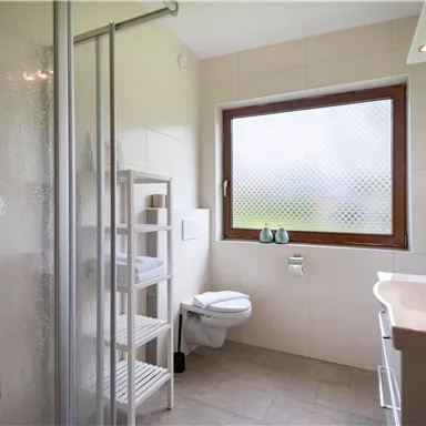 Ein modernes Badezimmer mit einer Dusche und einem Fenster. Es gibt ein Waschbecken, eine Toilette und einen Regal mit Handtüchern.