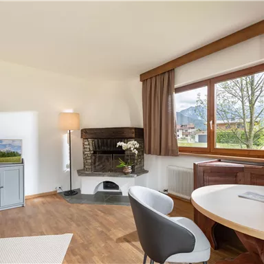 Ein gemütlicher Wohnraum mit einem Kamin, einem Tisch und modernen Möbeln. Große Fenster lassen viel Tageslicht herein und bieten einen Blick auf die Natur.