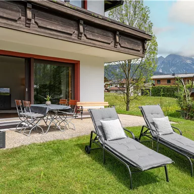 Ein modernes Wohnhaus mit einem gemütlichen Außenbereich. Zwei Liegen stehen im Gras, und im Hintergrund sind Berge zu sehen.