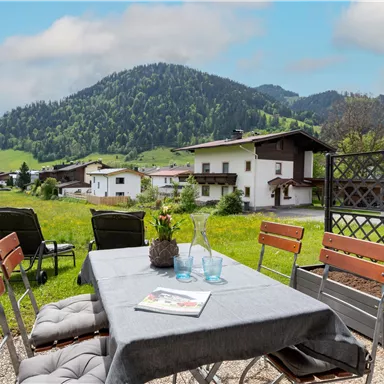 Ein gemütlicher Tisch im Freien mit Stühlen und Blick auf die Berge. Die Landschaft ist grün und hügelig, ideal für entspannte Stunden.