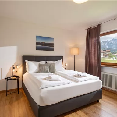 Ein gemütliches Schlafzimmer mit einem Doppelbett und einem Fenster mit Blick auf die Berge. Die warmen Holztöne und die sanfte Beleuchtung schaffen eine entspannende Atmosphäre.
