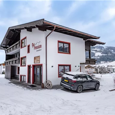 Ein modernes Haus im Schnee mit einem grauen Auto davor. Die Umgebung ist winterlich mit schneebedeckten Bergen im Hintergrund.