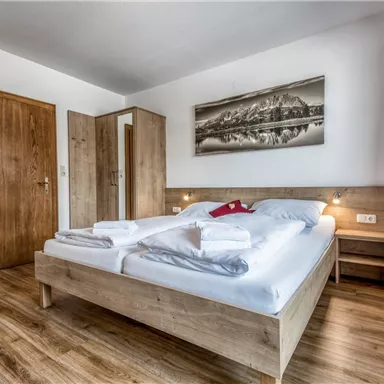 Ein modernes Schlafzimmer mit einem großen Doppelbett und Holzdekor. An der Wand hängt ein Landschaftsbild, und es gibt einen Schrank sowie einen Tisch.