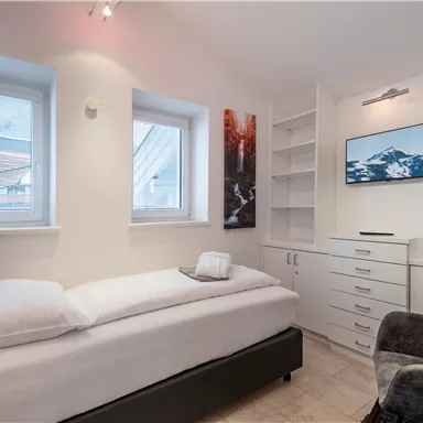 A small, modern bedroom with a single bed and a television on the wall. There are shelves and large windows that let in plenty of light.