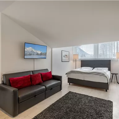 A modern bedroom with a double bed and a cozy sofa. There is a television on the wall and large windows that let in plenty of light.