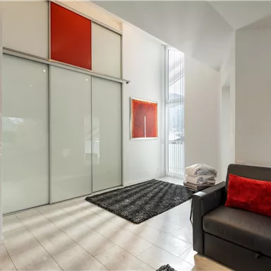 A modern living room with a gray sofa and red cushions. Large windows and a bright, minimalist decor characterize the space.