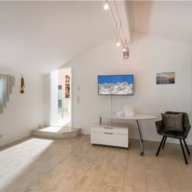 A modern room with slanted walls and hardwood flooring. There is a desk, a television, and large windows with horizontal blinds.