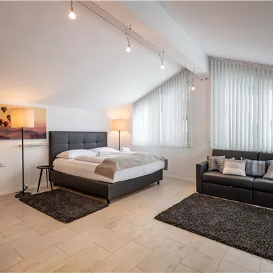A modern bedroom with a large bed, a sofa, and a bright, inviting atmosphere. Large windows provide plenty of daylight.