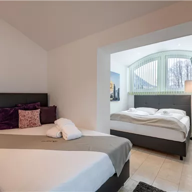 A modern bedroom with two beds and neutral colors. Large windows provide plenty of natural light.