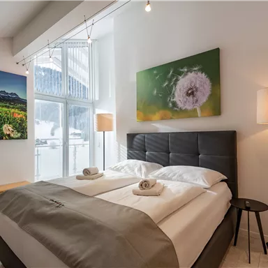 A bright bedroom with a double bed and two towels. Colorful pictures hang on the walls, and there is a large window front.