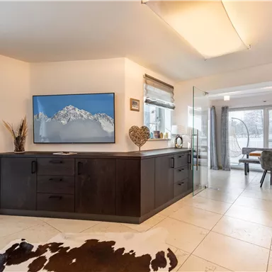 A modern living room with a large television and a view of the mountains. The floor is tiled with a rug, and there is a dining table in the background.