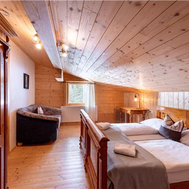 A cozy attic room with wooden paneling, two beds, and a small seating area. Large windows provide plenty of daylight and a view outside.