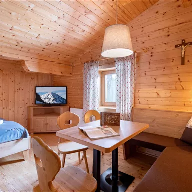 A cozy wooden room with a bed, a dining table, and a television. The walls are made of light wood, and a window lets in a lot of light.