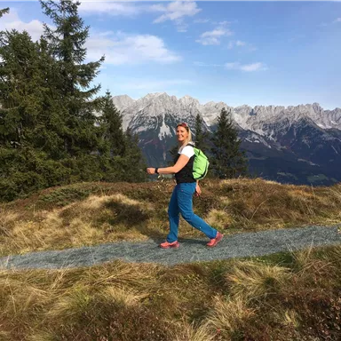 Eine Person wandert auf einem schmalen Weg in den Bergen. Im Hintergrund sind hohe Berge und ein blauer Himmel zu sehen.
