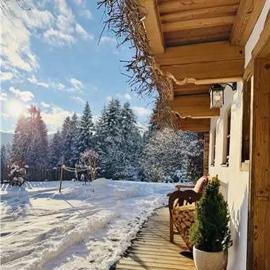 Eine gemütliche Terrasse eines Hauses im Schnee mit einem Blick auf schneebedeckte Bäume. Der Himmel ist klar und die Sonne scheint.
