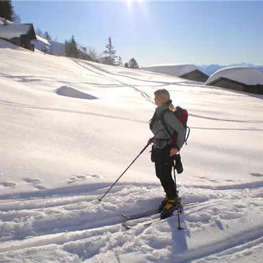 Eine Person steht auf einem verschneiten Hang und geht mit Skistöcken. Im Hintergrund sind schneebedeckte Hügel und Berghäuser zu sehen.
