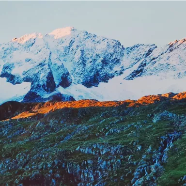 Eine beeindruckende Berglandschaft mit schneebedeckten Gipfeln und grünen Hängen. Die Morgenröte beleuchtet die Landschaft in warmen Farben.