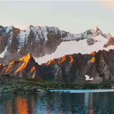 Eine beeindruckende Berglandschaft mit schneebedeckten Gipfeln und warmem Licht. Im Vordergrund spiegelt sich ein ruhiger See in den Bergen.